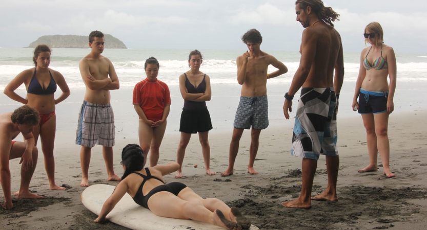A person demonstrates surfing skills on a beached board. Others stand around watching them. 